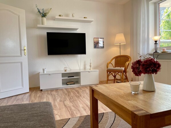 Un salon moderne avec un téléviseur à écran plat fixé au mur, une table à manger en bois, un fauteuil en osier et un vase rempli de fleurs.