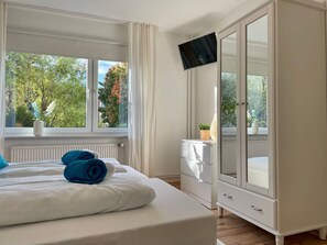 A hotel room with a bed, a TV mounted on the wall, a window with a view of trees, and a white wardrobe. - Haus Jenina - House Jenina - Directly on the dike, modern vacation apartment for up to 6 persons (Cuxhaven)