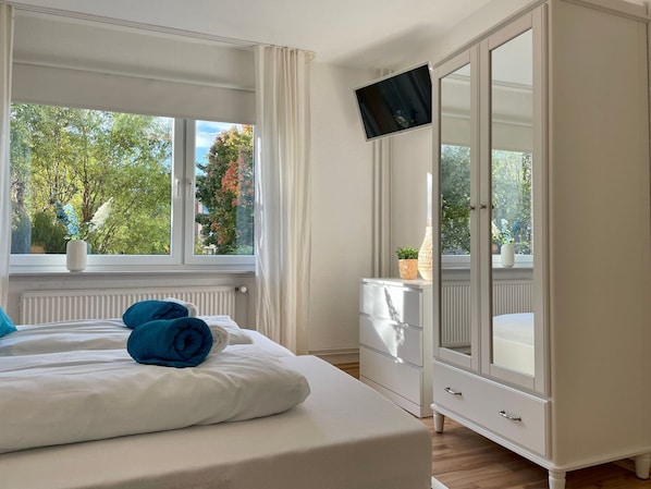 A hotel room with a bed, a TV mounted on the wall, a window with a view of trees, and a white wardrobe.