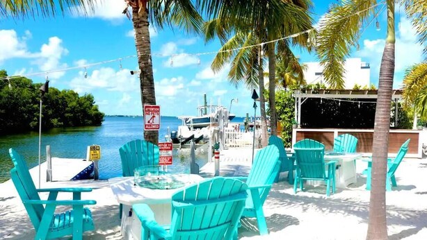 Un espacio de descanso frente a la playa con sillas turquesas y una mesa, con vistas a un puerto deportivo lleno de barcos.