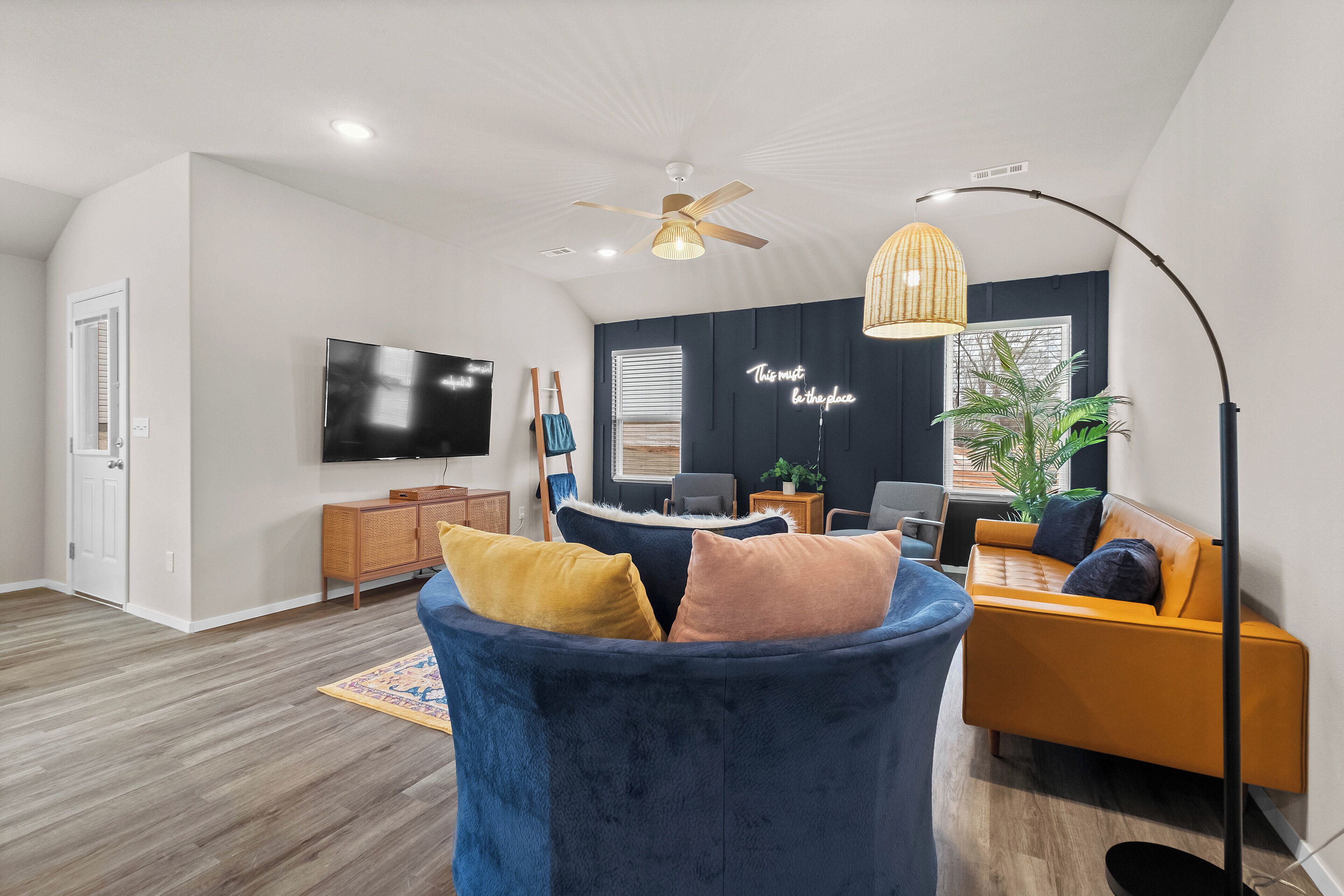 A modern living room with a dark blue sofa, a yellow and pink pillow, a wooden cabinet, a television, and a ceiling fan.