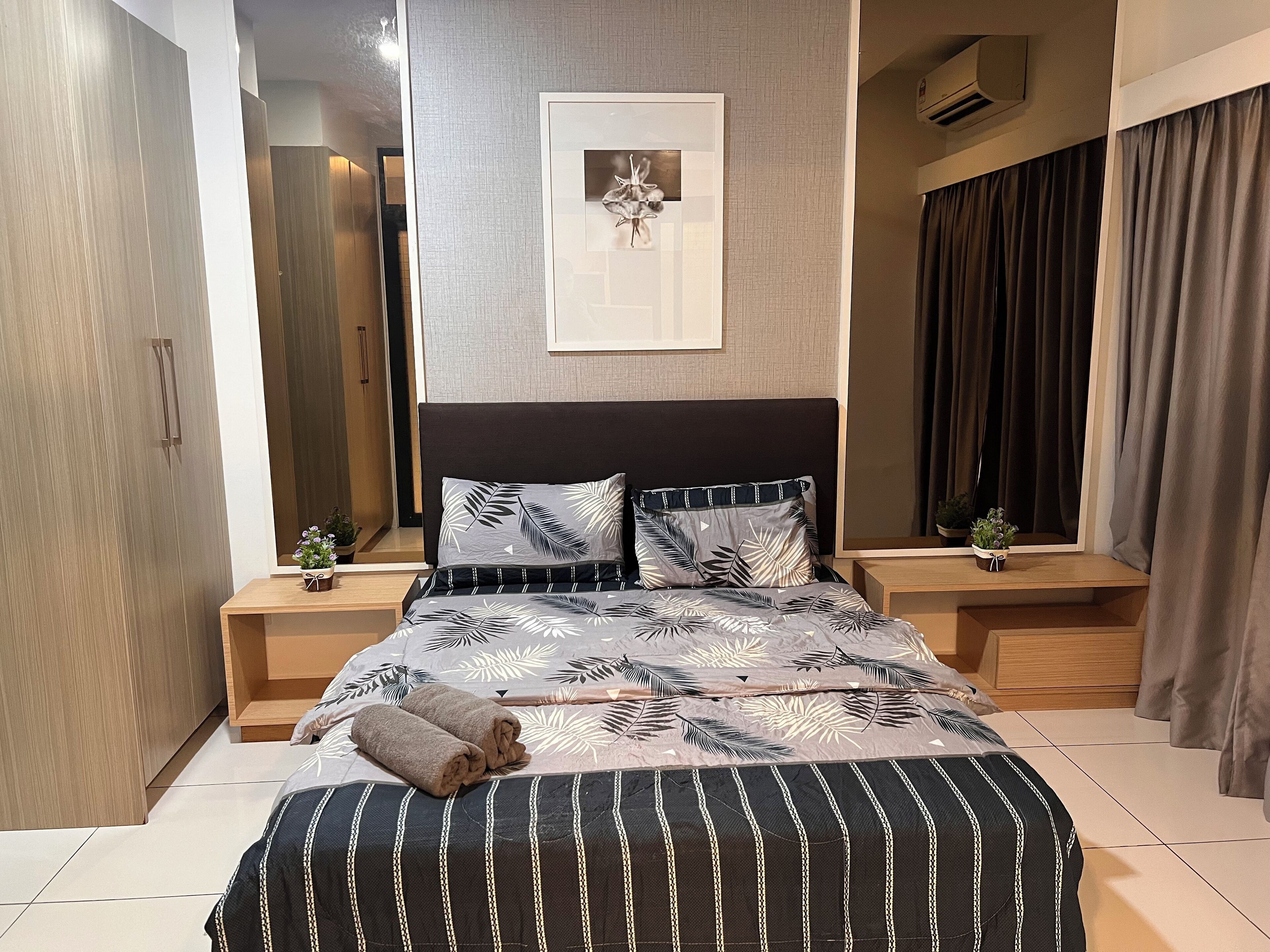 A modern bedroom with a large bed, a headboard with a decorative mirror, bedside tables with small plants, and a view of the outside through a window with curtains.