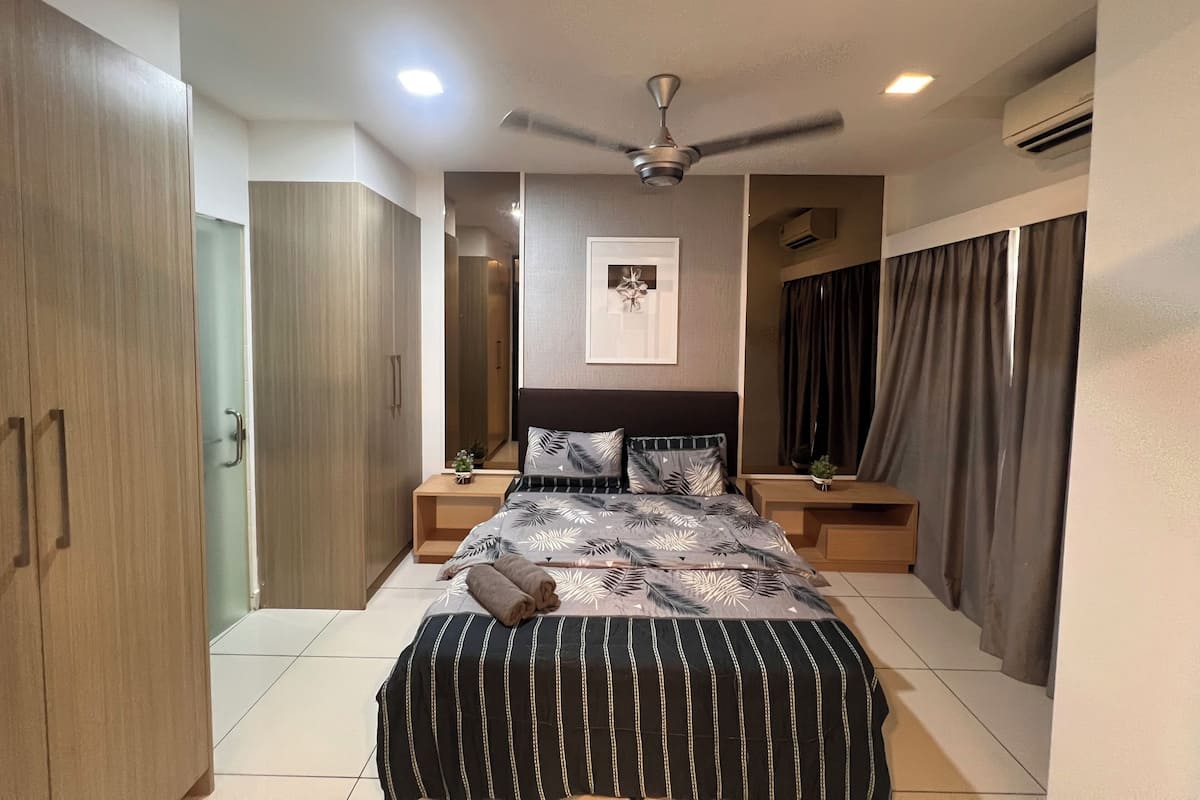 Quarto com cama, mesas de cabeceira, ventoinha no teto, espelho e janela com cortinas.