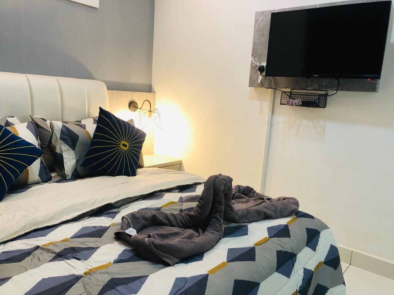 A neatly made bed with a geometric patterned bedspread, a white headboard, and decorative pillows. A flat-screen TV is mounted on the wall above the bed.