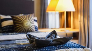 A neatly made bed with a blue and white patterned bedspread, a dark blue pillow, and a decorative pillow with a sunburst design. A lamp with a white shade is on a bedside table, and a curtain is partially visible. - 1 Tebrau Residences by YML Home (Johor Bahru)