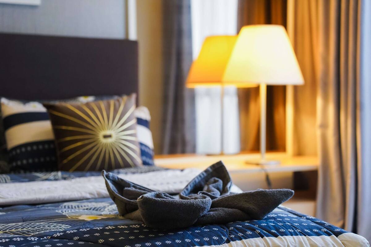 Uma cama bem arrumada com um cobre-leito estampado em azul e branco, um travesseiro azul escuro e um travesseiro decorativo com padrão de raios de sol. Uma luminária com cúpula branca está sobre uma mesa de cabeceira, e uma cortina aparece parcialmente.