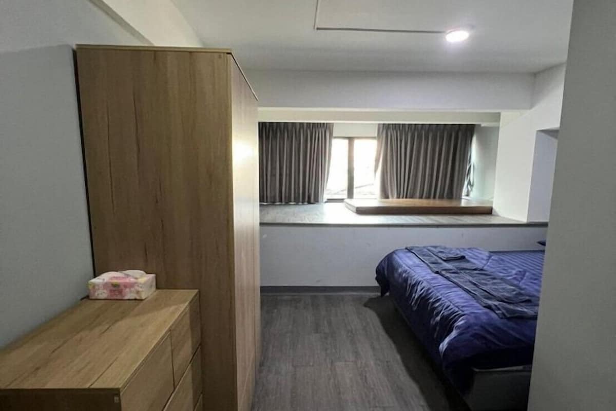 Ein schmales Zimmer mit einem hölzernen Schminktisch, einem Bett mit blauer Bettdecke und einem Fenster mit Vorhängen.