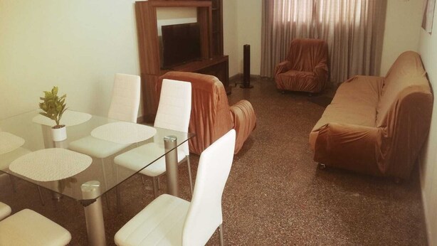 Una sala de estar con una mesa de comedor de vidrio, sillas blancas, un sofá marrón, un sillón marrón y un televisor sobre un mueble de madera.