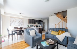A modern living room with a grey sofa, a coffee table, and a dining area with a wooden table and chairs. A kitchen with dark cabinets and a wooden staircase is visible in the background.