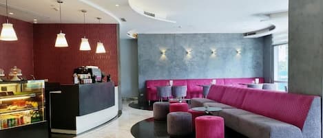 A modern hotel lobby with a curved reception desk, a seating area with a sofa and ottomans, and a food display.