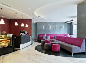 Un moderno vestíbulo de hotel con un mostrador de recepción curvo, una zona de estar con sofá y pufs, y una presentación de alimentos.
