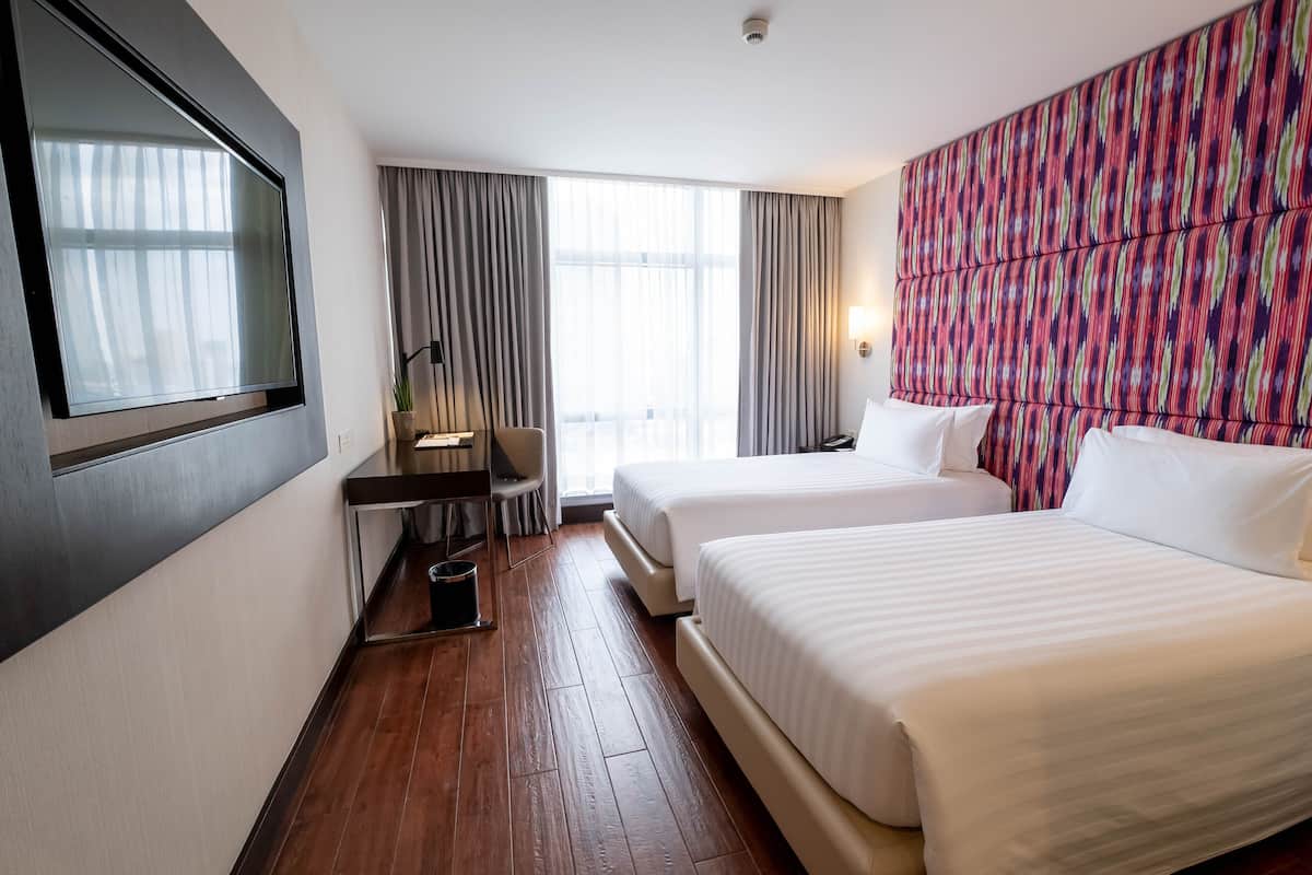 A hotel room with two beds, a desk, a chair, and a large window with curtains.