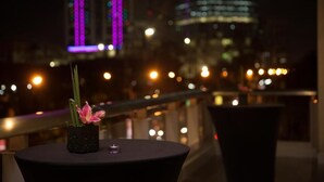 Vista nocturna desde un balcón con un florero y una vela sobre una mesa, con vistas a un paisaje urbano con edificios iluminados.