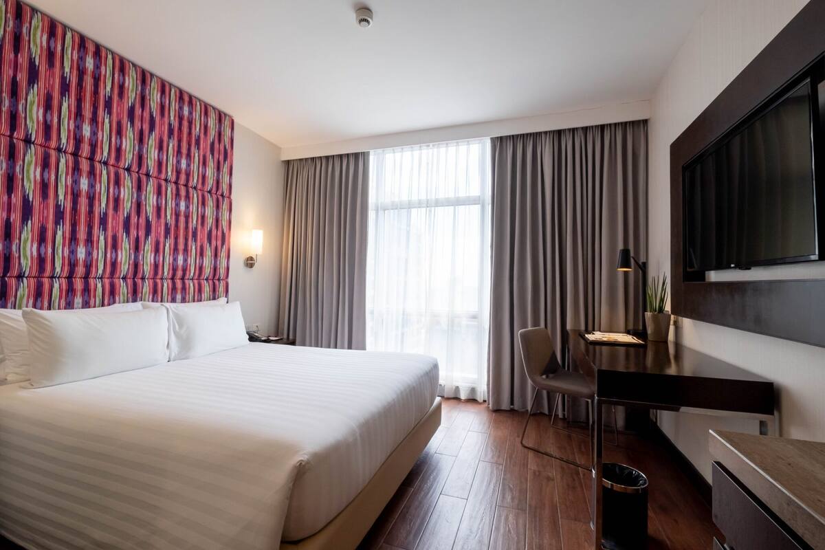 A hotel room with a large bed, a desk with a chair, a TV mounted on the wall, and a window with curtains.