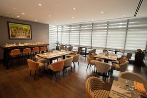 A dining area with multiple tables and chairs, large windows with blinds, and a painting on the wall.