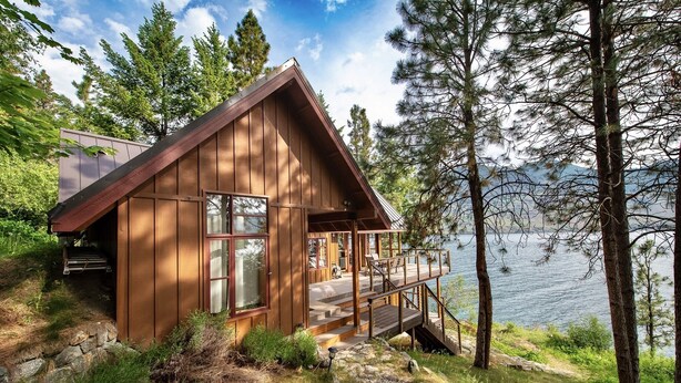 Eine Holzhütte mit einer Terrasse, die auf einen See mit umliegenden Bäumen blickt.