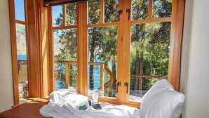 Ein Zimmer mit großem Fenster, einem Bett mit weißer Bettwäsche und Blick auf Bäume und ein Gewässer.