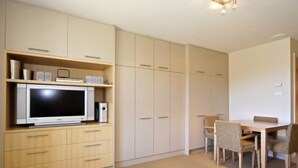 Un espace de vie compact comprenant un téléviseur posé sur un meuble en bois, une table à manger avec des chaises et des placards intégrés.