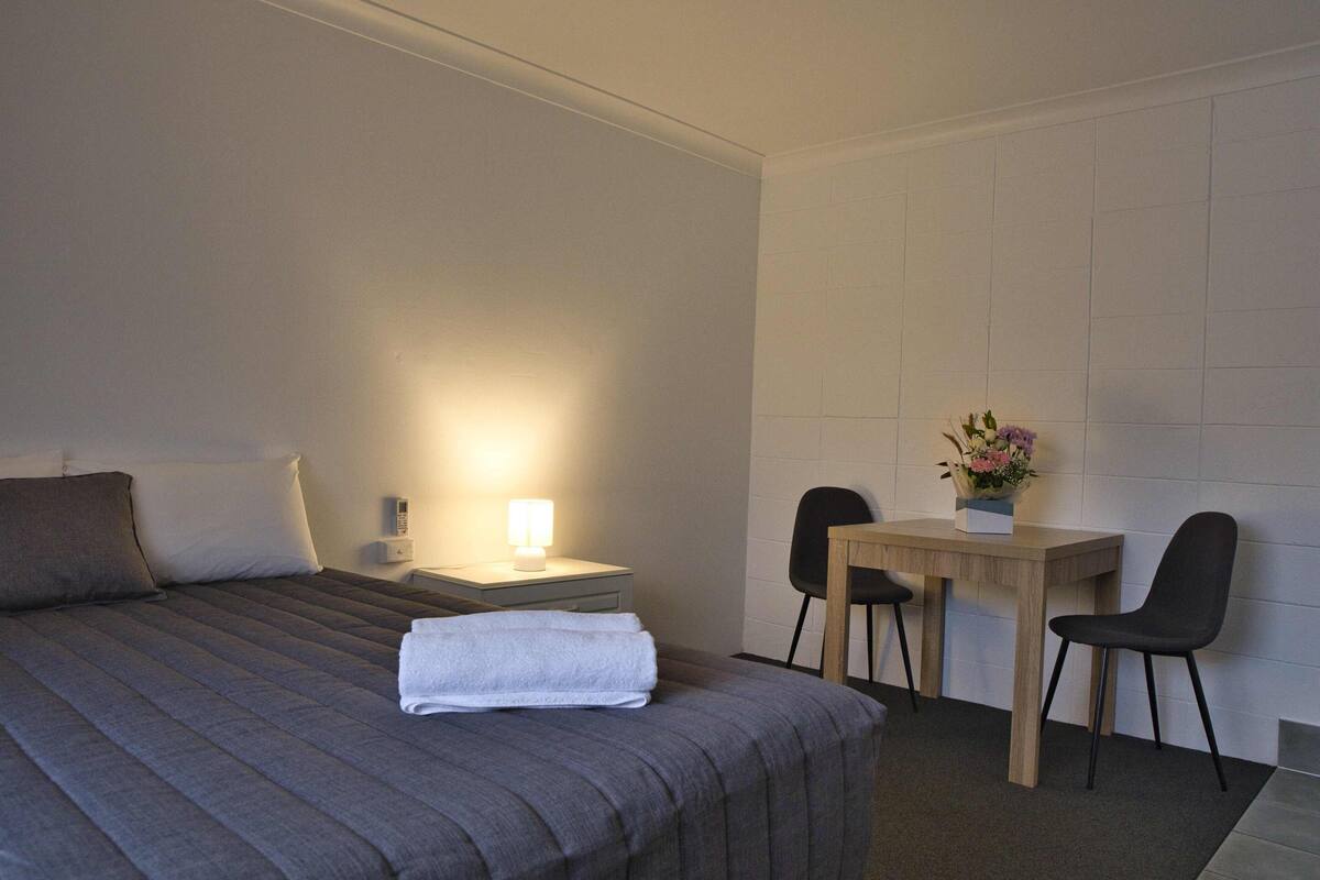 A hotel room with a bed, bedside table, lamp, and a small table with two chairs and a vase of flowers.
