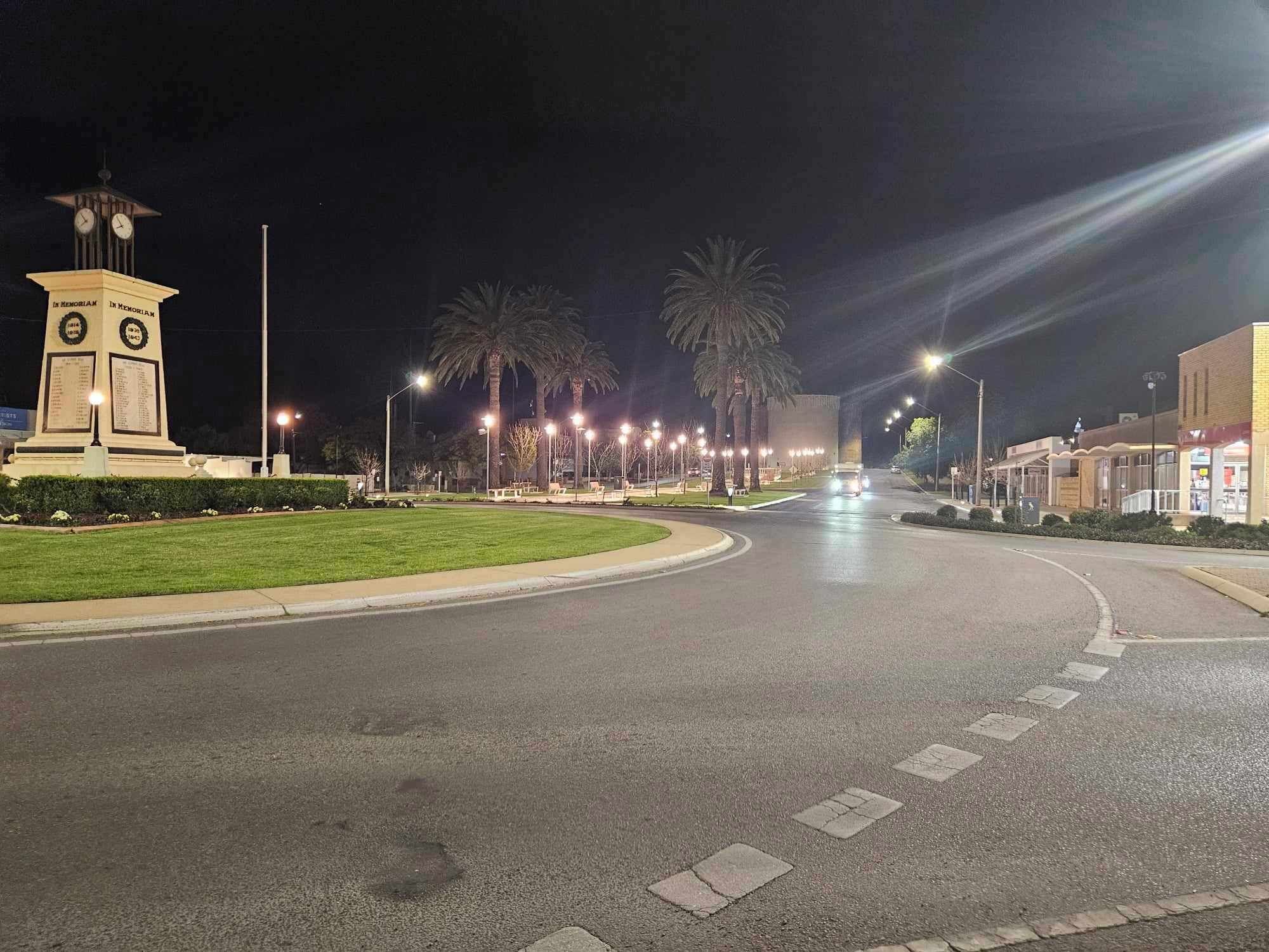 Calle bien iluminada de noche con un monumento que presenta un reloj y placas, palmeras y una carretera curva que lleva a un edificio con tiendas.