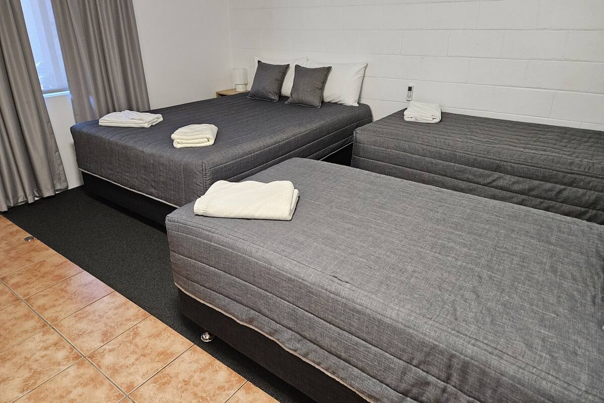 A hotel room with two single beds, each with a grey mattress and pillows, and a small bedside table with a lamp.