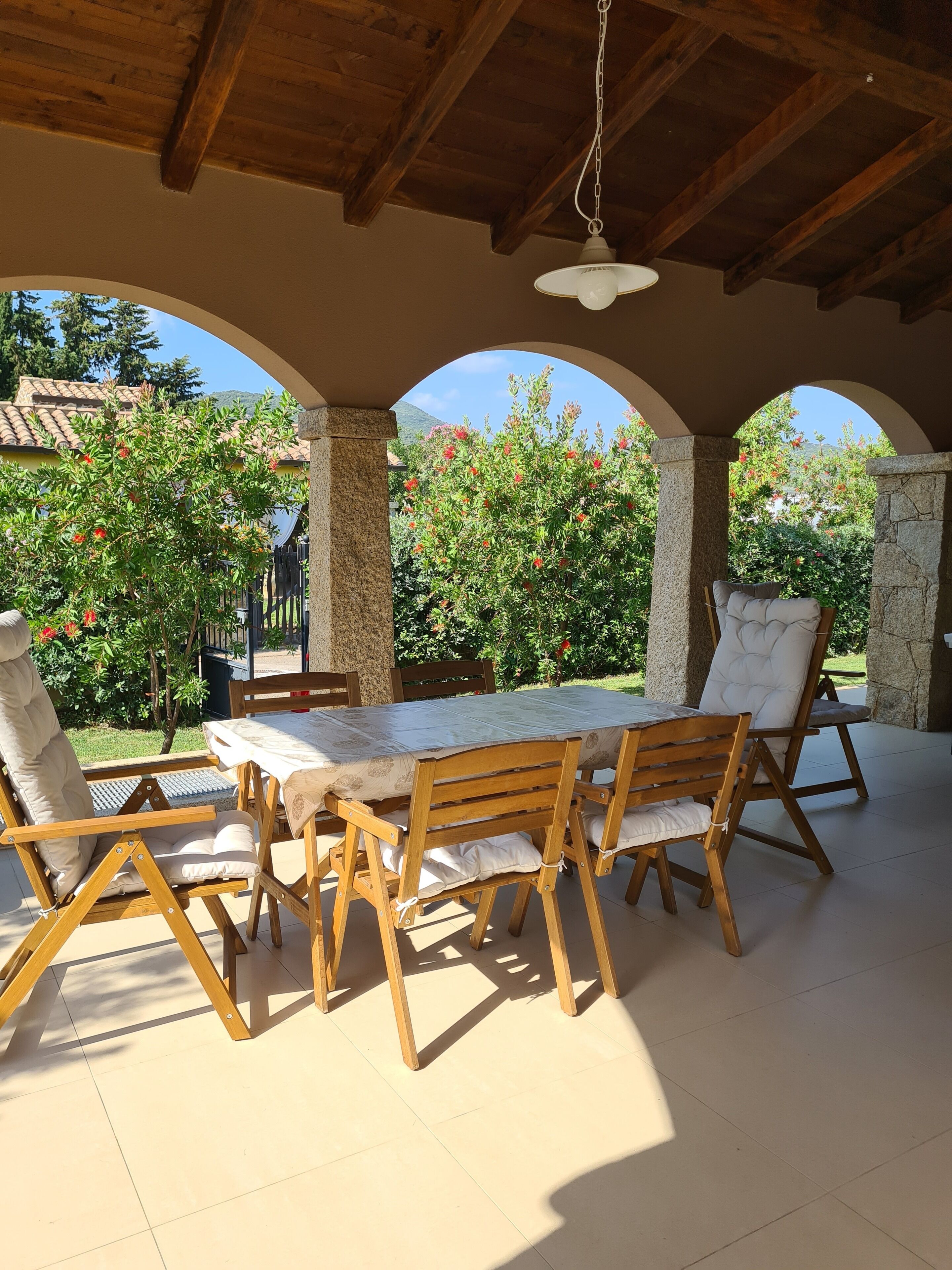 Un patio esterno con tavolo e sedie di legno, pilastri in pietra e vista su alberi e palazzi.