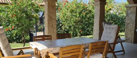 Un patio esterno con tavolo e sedie di legno, pilastri in pietra e vista su alberi e palazzi.