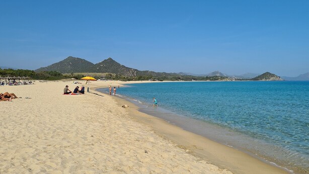 Une plage à l’eau d’un bleu limpide, bordée d’un rivage sablonneux, avec des montagnes qui se dessinent à l’horizon.