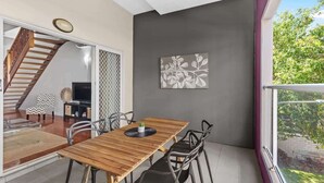 A dining area with a wooden table and chairs, a TV on a stand, a staircase, and a balcony with a view of greenery. - Hipster Loft! ~  1Bed/Study/1Bath/Balconies/1Car (Fortitude Valley)