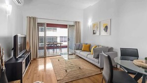 A living room with a sofa, TV, glass table, and a view through a sliding door to an outdoor balcony. - Hipster Loft! ~  1Bed/Study/1Bath/Balconies/1Car (Fortitude Valley)
