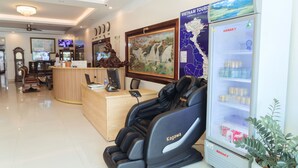A reception area with a massage chair, a refrigerator stocked with beverages, a wooden desk, and a wall-mounted television. - Luxury Airport Hotel & Travel (Hanoi)