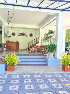 L’entrée d’un hôtel, avec un escalier menant au hall d’accueil, un comptoir de réception et une enseigne indiquant « HÔTEL EDEN GRAND ».