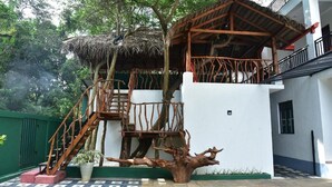 A treehouse structure with a wooden staircase and a thatched roof, attached to a white building with a balcony. - Eden Grand (Dambulla)