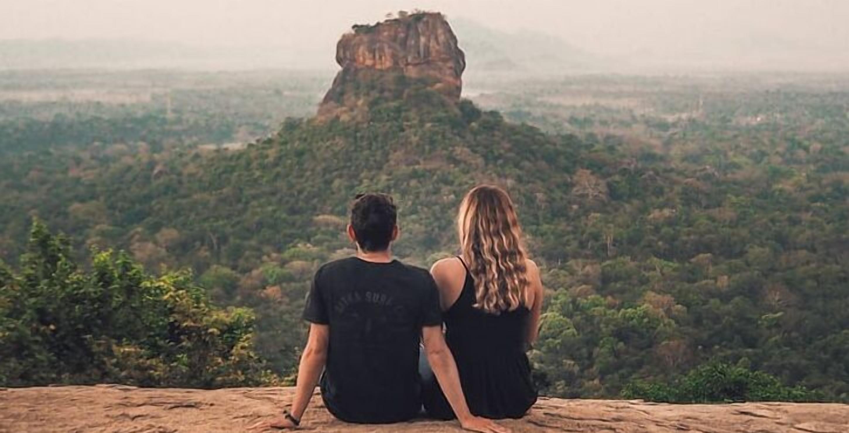 Zwei Personen sitzen auf einem Felsen und blicken auf eine gebirgige Landschaft.
