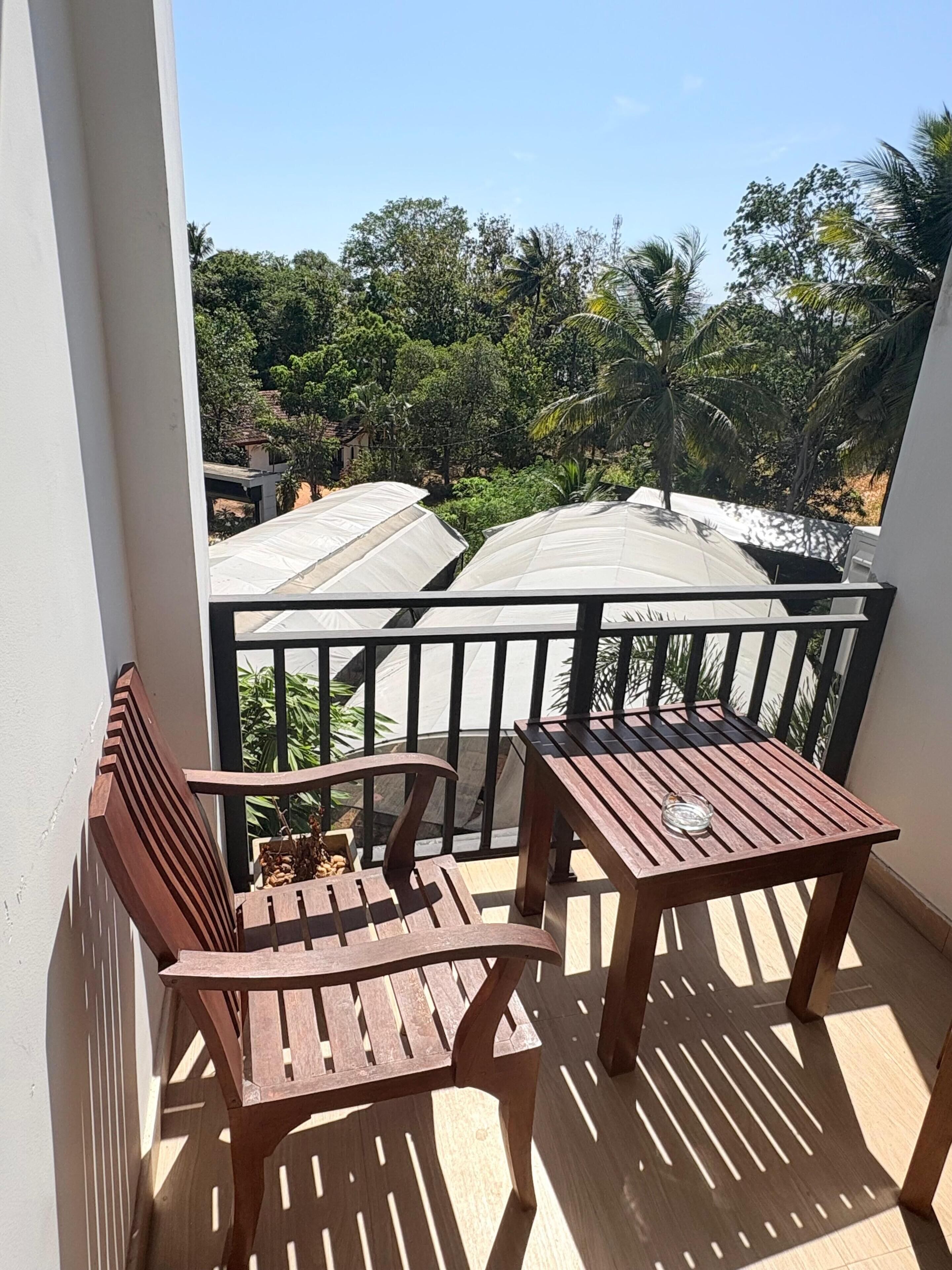 A balcony with a wooden table and chairs, a view of a residential area with trees and houses, and a clear sky.
