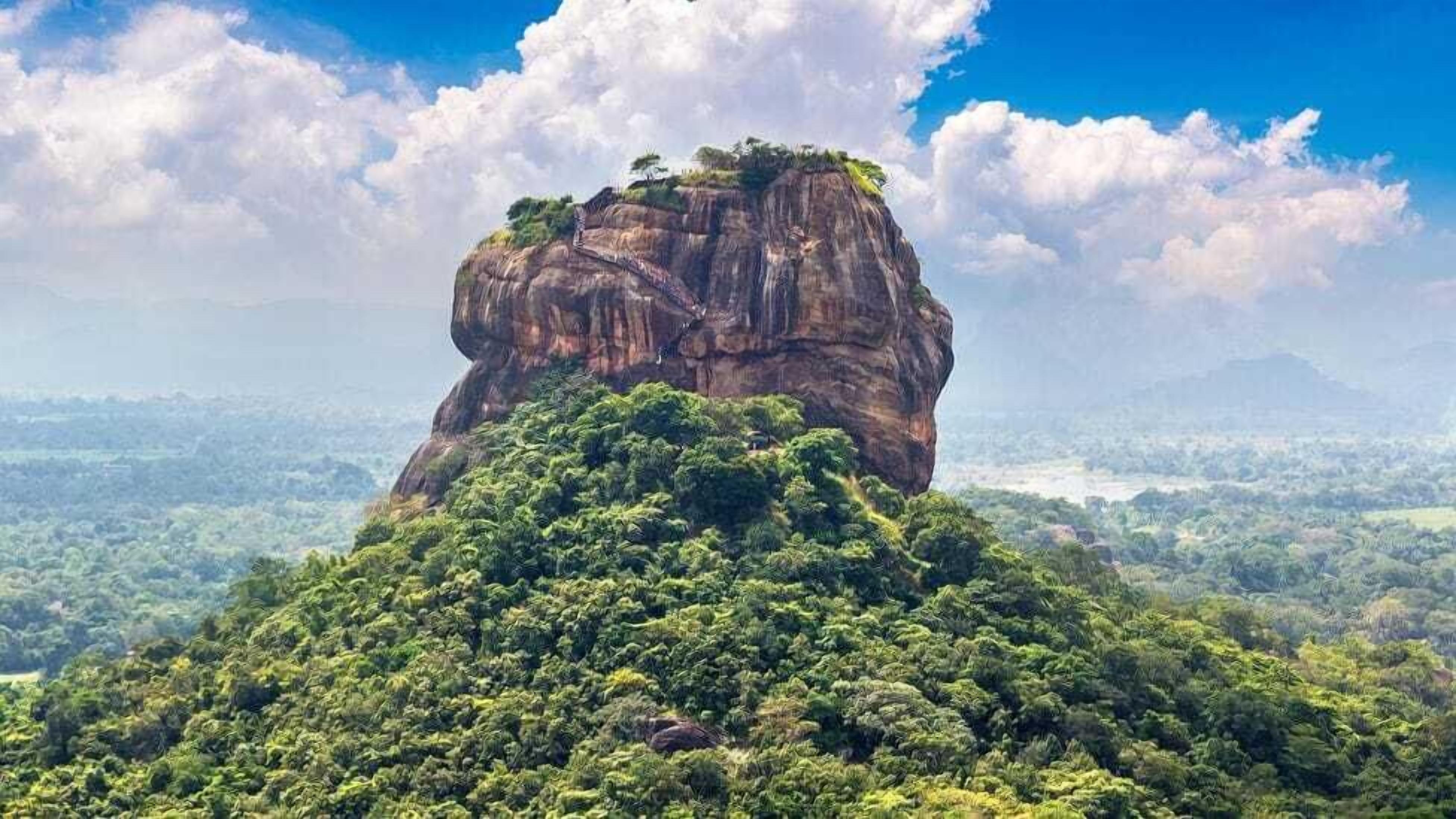 A large rock formation with dense green vegetation at its base, surrounded by a vast landscape.