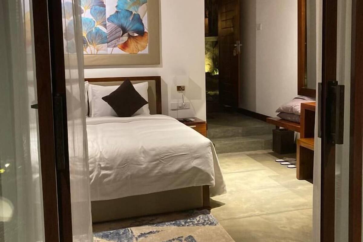 A bedroom with a bed, a ceiling fan, a painting, and a view into another room.