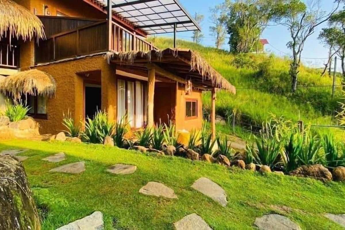 A two-story house with a thatched roof, a wooden balcony, and a stone pathway leading to the entrance.