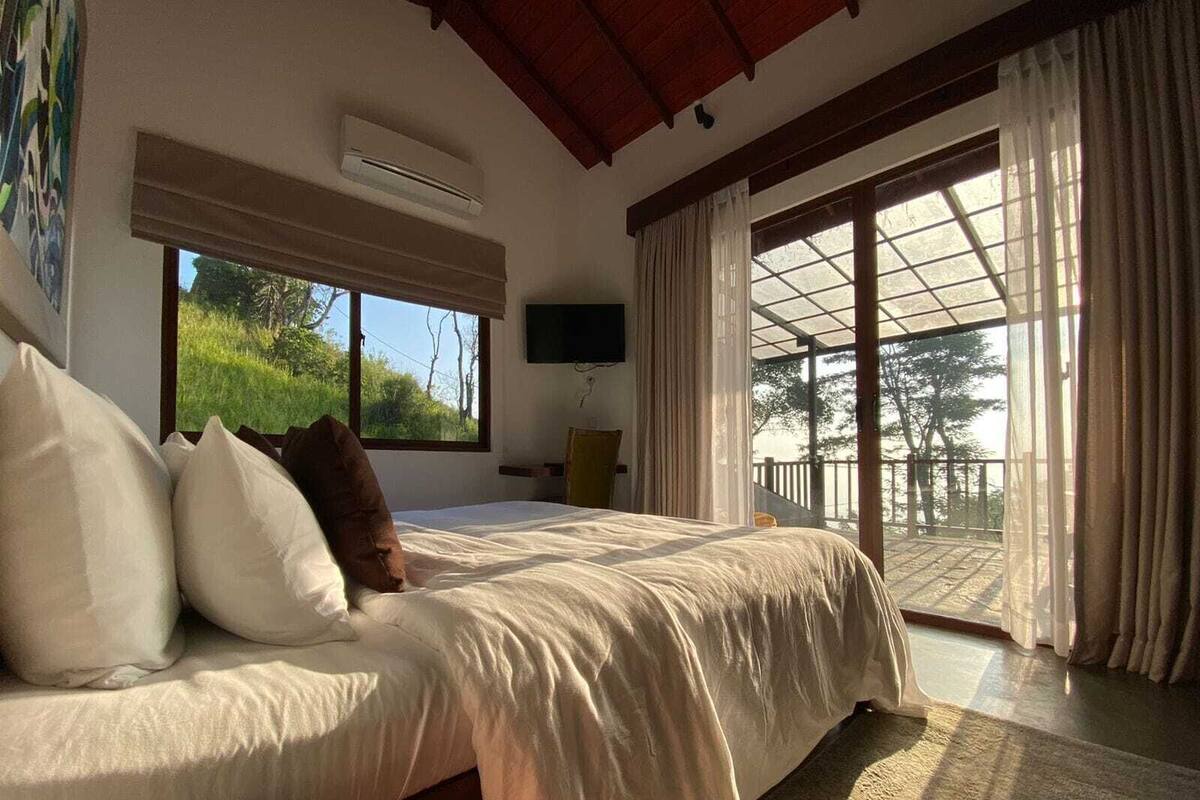 A bedroom with a bed, a TV, a large window, and a balcony.