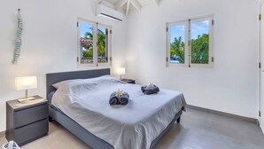 Un dormitorio con una cama, dos mesitas de noche, una lámpara y dos ventanas con vistas a palmeras y un edificio.