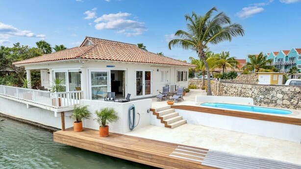 Una propiedad frente al agua con un porche cubierto, mobiliario exterior, piscina y un muro de piedra.
