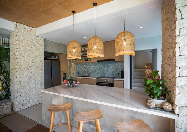 A modern kitchen with a large island, wooden bar stools, and hanging wicker pendant lights.