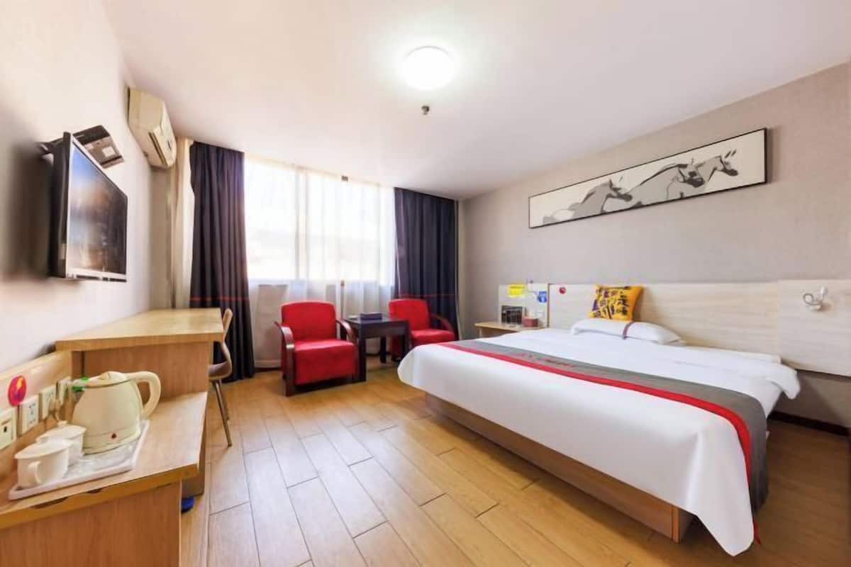 Habitación de hotel con cama, escritorio con hervidor, dos sillas rojas, televisión y ventana con cortinas.