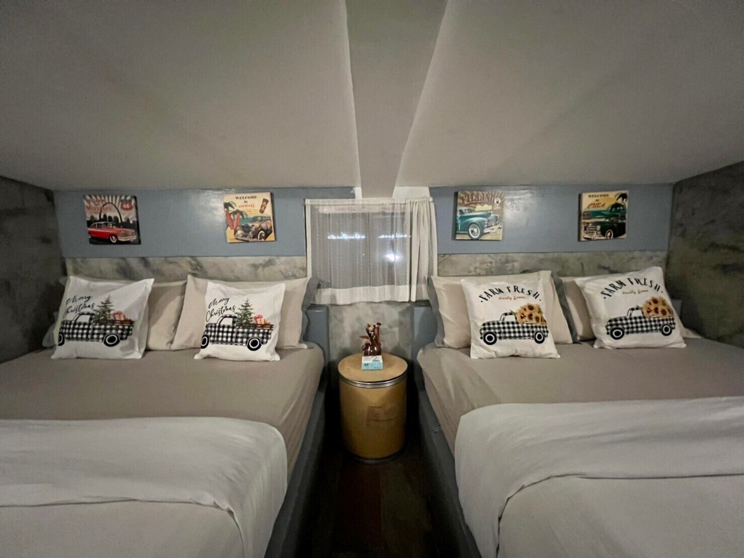 A room with two beds, each with a pillow featuring a car design. There are wall decorations, a small round table with a candle, and a window with sheer curtains.