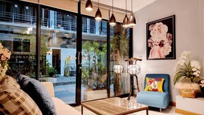 A modern living room with a sofa, a blue armchair, a coffee table, and a large window with a view of a building and greenery outside. - Baan Bua Talay (Rayong)