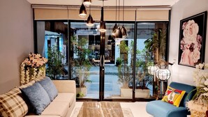 A modern living room with a sofa, a blue armchair, and a large glass door leading to an outdoor area with plants and a view of a building. - Baan Bua Talay (Rayong)