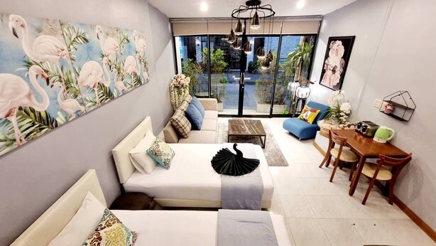 A modern living room with a sofa, a dining table, and a coffee table. There is a painting of flamingos on the wall and a view of the outside through large windows. - Baan Bua Talay (Rayong)