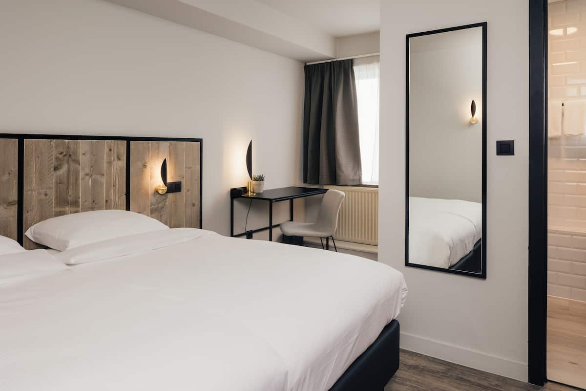 Quarto de hotel moderno com uma cama grande, uma escrivaninha, uma cadeira e um espelho.