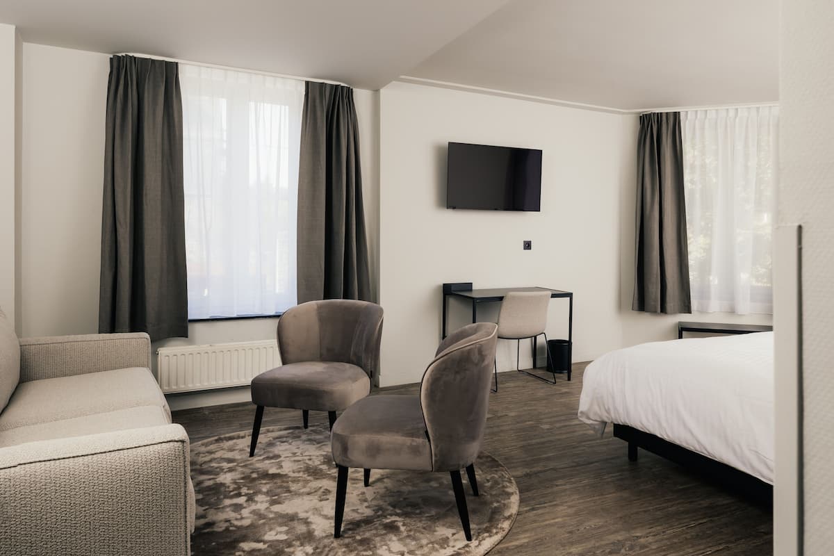 Quarto de hotel com cama, duas poltronas, escrivaninha, televisão e janelas amplas com cortinas.