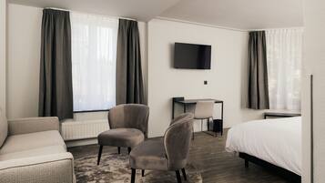 Une chambre d’hôtel comprenant un lit, deux fauteuils, un bureau, une télévision et de grandes fenêtres avec des rideaux.